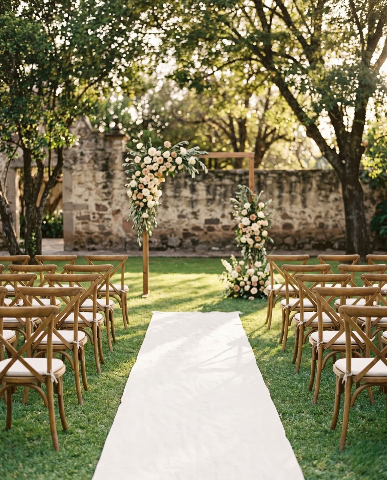 Ceremonia de boda en jardín: pasillo de lino blanco entre sillas de madera, hacia un arco de flores en tonos crema junto a un muro de piedra